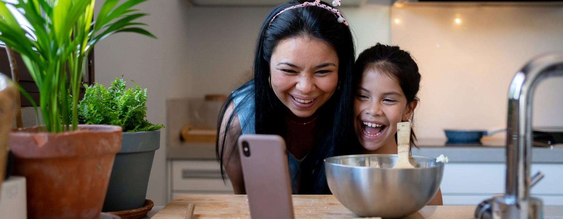 mother and young daughter smile at their phone as they cook in the kitchen